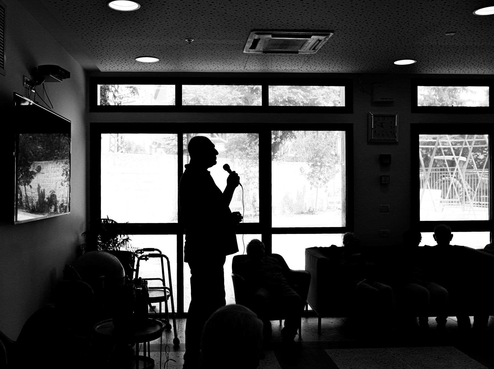 A speaker silhouetted against a window while a small audience listens